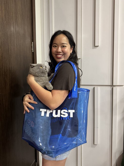A smiling young woman holds a gray British Shorthair cat while wearing a blue tote bag labeled 'TRUST' and a lanyard reading 'Virtual Travel Agent'.