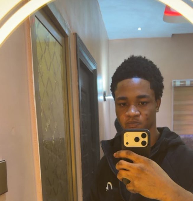 A person wearing a black hoodie takes a mirror selfie with a smartphone in a warmly lit hallway.