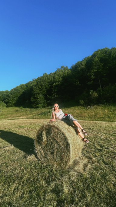 A woman in a white top and shorts sits on a large round hay bale in a grassy field with trees and a clear blue sky in the background.
