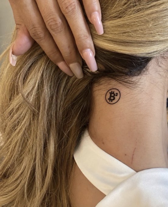A close-up of a small Bitcoin symbol tattoo on the side of a person's neck, with blonde hair pulled back and a hand resting near the head.