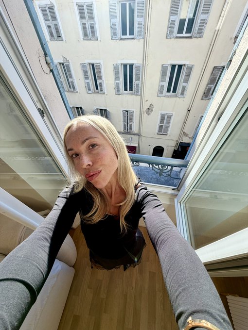 A blonde woman in a black top takes a selfie from a high angle, with her arms extended toward the camera, overlooking a European-style courtyard with shuttered windows.