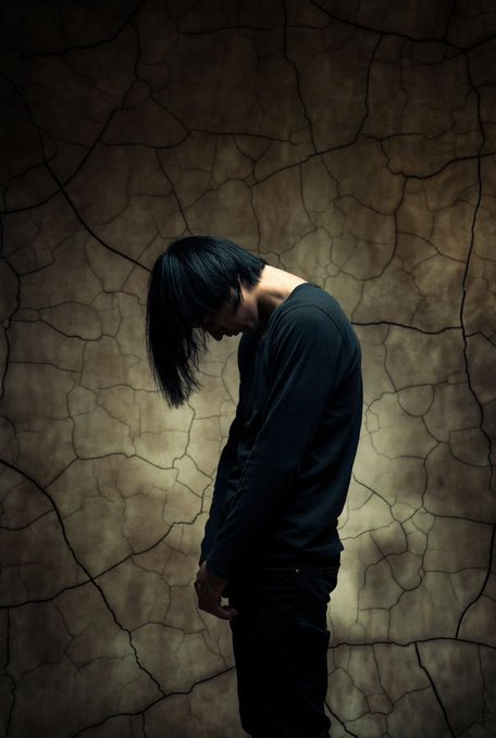 A dark, moody portrait of a person with long black hair looking downward against a textured, cracked brown background.
