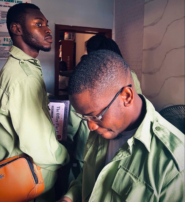 Two young men wearing matching olive-green jackets stand in what appears to be a small room or shop, with one looking down wearing glasses while the other gazes off to the side.