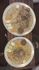 Two large plates of biryani rice topped with meat, sliced boiled eggs, cucumbers, and carrots served on a wooden table.