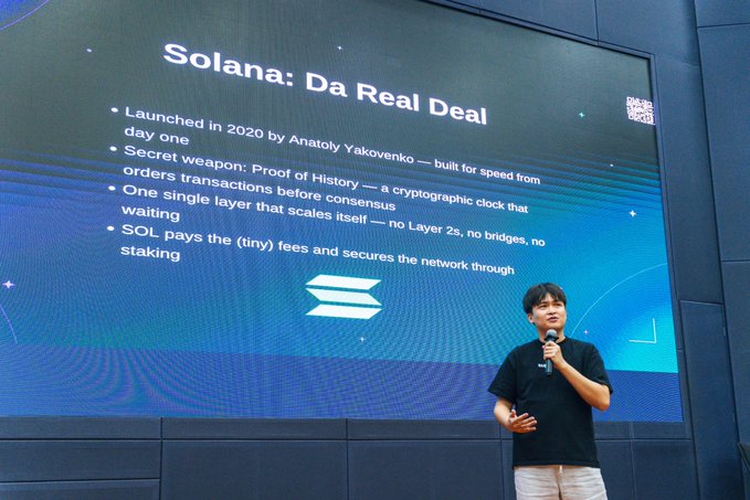 A presenter speaking on stage in front of a large screen titled 'Solana: Da Real Deal', which highlights Solana's 2020 launch by Anatoly Yakovenko and its Proof of History cryptographic clock.