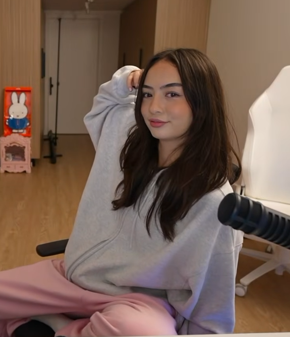 A young woman poses in a gray zip-up hoodie and pink pants, smiling at the camera in a softly lit room with a Miffy figurine visible in the background. Her casual, cute aesthetic matches the post's simple compliment "qt" (short for "cutie").