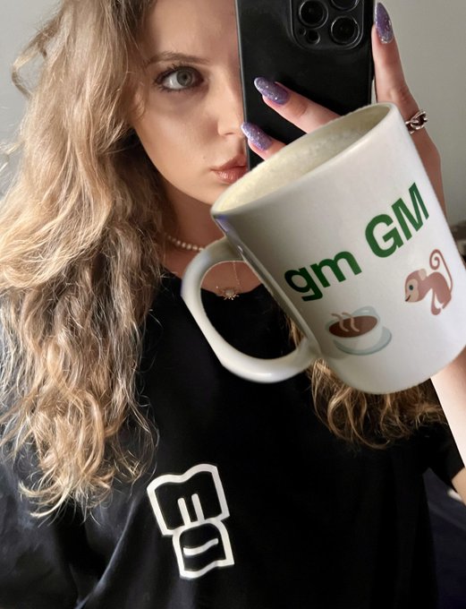 A blonde woman takes a mirror selfie with a phone, wearing a black shirt and holding a white mug that reads 'gm GM' with a squirrel and coffee design.
