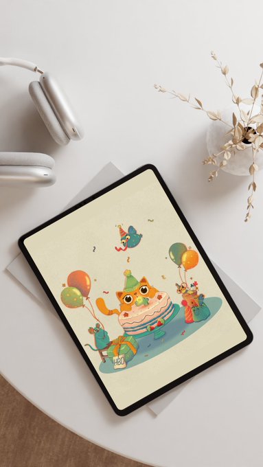 A flat-lay photograph shows a tablet displaying a whimsical birthday illustration of a cat-shaped cake with a party hat surrounded by balloons and animals, styled with white headphones and dried pampas grass on a white surface.
