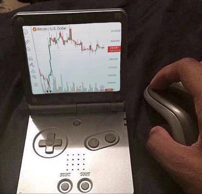 A Nintendo Game Boy Advance SP displaying a Bitcoin/U.S. Dollar price chart showing BTC at 29287, held next to a person's hand using a computer mouse.