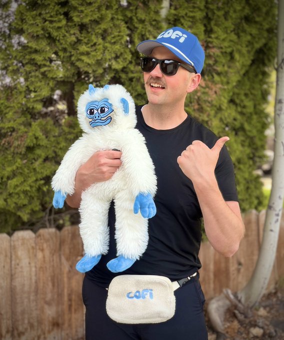 A man wearing a "LOFI" branded hat and sunglasses smiles while holding up a blue and white yeti plush toy and giving a thumbs-up, with a matching "LOFI" fanny pack around his waist. The image visually matches the post's caption about embracing the $LOFI memecoin, featuring the mascot plushie as a promotional nod to @lofitheyeti.