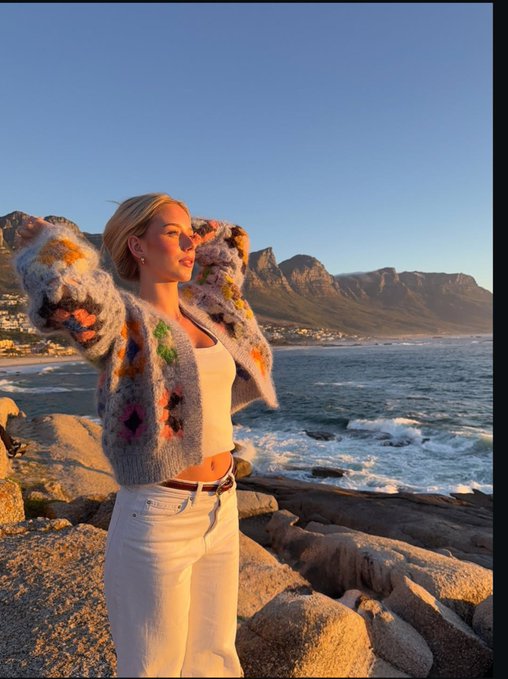 A young woman poses on rocky coastal terrain at sunrise, wearing a colorful patterned fuzzy jacket, white tank top, and white jeans, with mountains and ocean behind her. The "Good Morning Friends, Can I Get GM Back?" caption pairs naturally with her peaceful, sunlit morning greeting on the scenic shore.