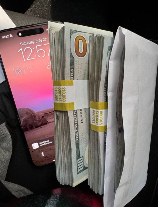 A smartphone displaying the time 12:57 on Saturday, July 27 rests next to a thick envelope stuffed with banded stacks of $100 bills totaling over $10,000 per band, visually reinforcing the caption "Money must be made."