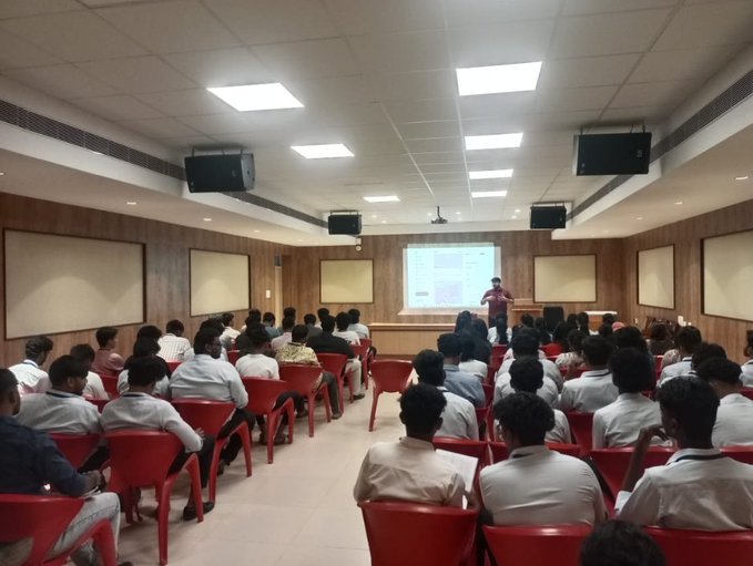 A lecture hall filled with students in white shirts sitting in red chairs facing a presenter at the front near a projection screen.