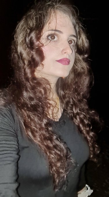 A portrait photo of a woman with long, curly brown hair and red lipstick against a dark background.