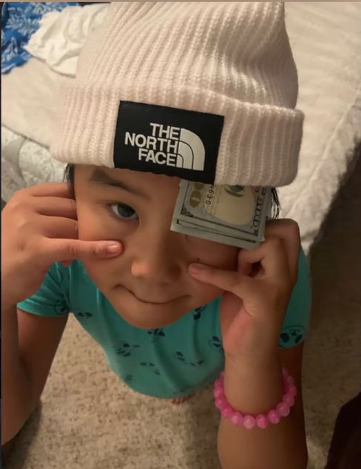 A child wearing a white 'The North Face' beanie and a teal shirt, holding a folded US dollar bill over one eye while posing playfully for a selfie.