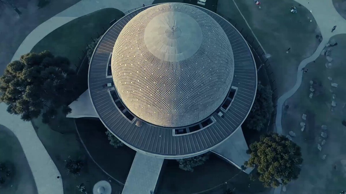 The image features an aerial view of a large dome-shaped building with a unique design. The building appears to be a museum or a significant landmark within its surroundings. There are several people visible around the area, possibly admiring the structure or going about their daily activities. A clock can also be seen in the scene, indicating the time for those present at the location.