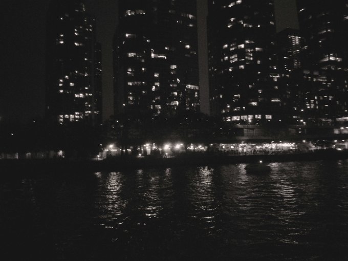 A black and white nighttime photograph showing city skyscrapers with illuminated windows reflected in a body of water in the foreground.