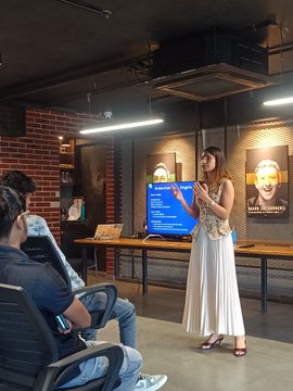 A woman in a white pleated skirt and beige top stands presenting in front of a large screen displaying charts, while a man in a gray shirt and cap sits watching in an office space.