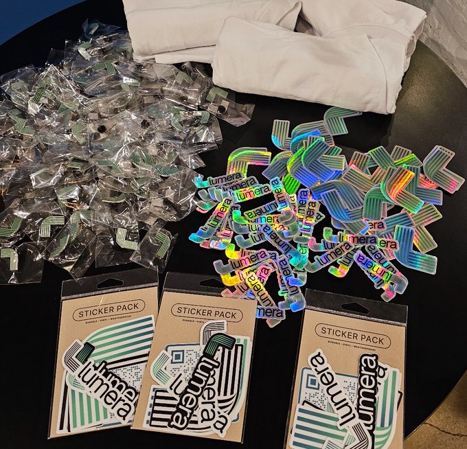 A collection of merchandise on a black table, including individually wrapped holographic stickers, a pile of holographic stickers reading "lúmera", and three sticker packs labeled "STICKER PACK" with the "lúmera" logo, alongside folded white clothing.