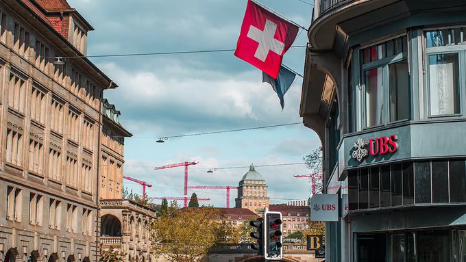 A street view in Switzerland featuring a prominent Swiss flag and UBS bank signage on a building, with historic architecture, construction cranes, and a domed building visible in the background.