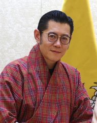 A man in glasses wearing a traditional checkered patterned robe (likely a Bhutanese "gho") stands in front of a yellow flag, with the post's Bitcoin mining reference connecting to Bhutan's well-known use of its hydroelectric power for Bitcoin mining operations.
