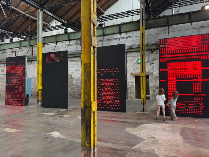 An industrial warehouse space with yellow support columns displays several large black canvases featuring red text-based artwork, with two visitors examining one of the pieces.