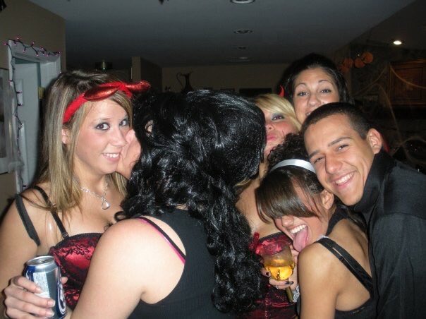A group of young people at a party, with some wearing costumes including devil horns and black wigs, holding drinks.