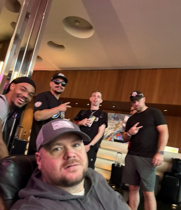A selfie-style group photo of five men, including one in the foreground wearing a gray hoodie and cap, and others in the background posing with hand gestures in what looks like a hotel lobby.
