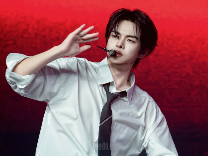 The image features a young man wearing a white shirt, black tie, and holding a microphone close to his mouth with one hand while waving the other hand outwards. The background of the photo has red hues that contrast with the white shirt and tie, making them stand out. The caption "260620 SMA #리오 파드라이브원 합니다" is also present in the image.