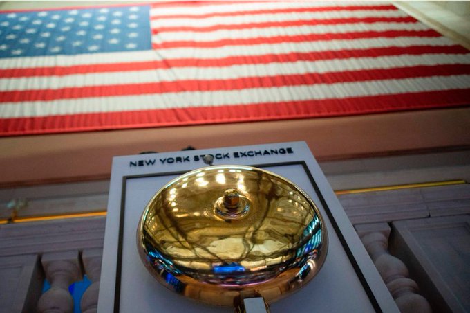 A close-up of a golden New York Stock Exchange bell mounted on a plaque, with an American flag displayed in the background.