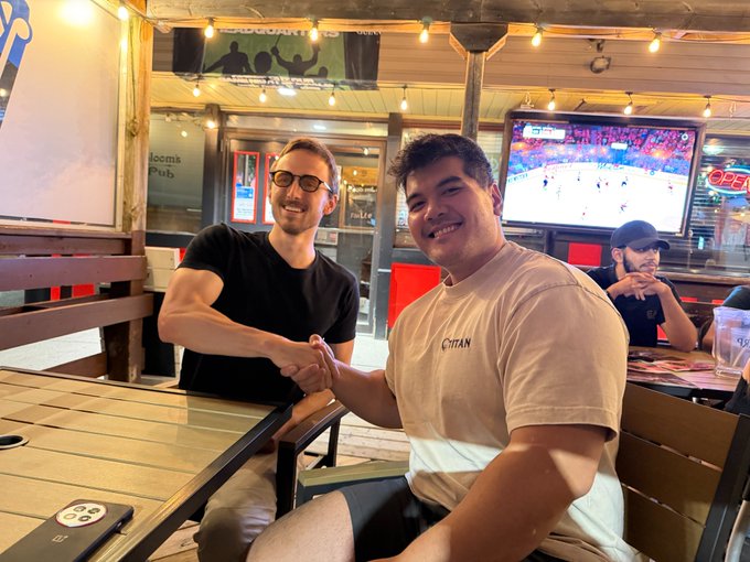 Two men shake hands and smile at each other across a table in what appears to be an outdoor restaurant setting, with a TV showing a sports game in the background.