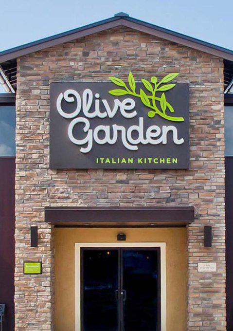 The image shows the exterior entrance of an Olive Garden Italian Kitchen restaurant, featuring its signature green and white logo with an olive branch design mounted on a stone facade above the glass doors. The image relates to the post's playful timeline scenario, humorously framing the Olive Garden storefront as the backdrop for an NFT "soulbound" pass dutch auction.