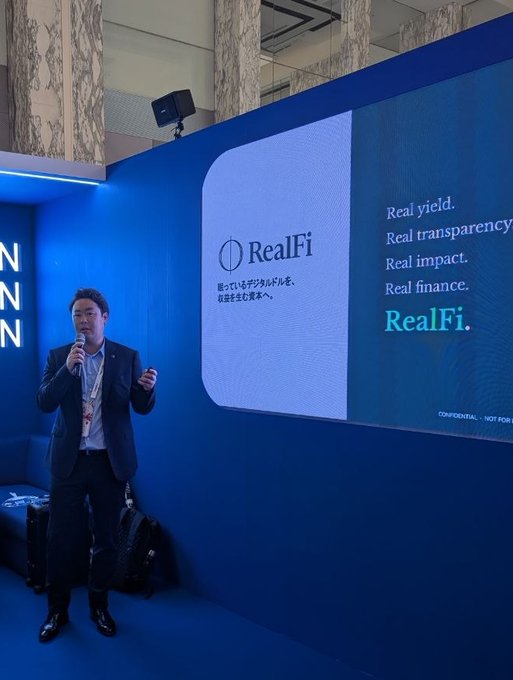 A man in a suit speaks into a microphone while presenting "RealFi" at the Cardano booth, with a large screen behind him displaying the RealFi logo alongside the tagline "Real yield. Real transparency. Real impact. Real finance. RealFi." The image captures the moment shared in the post about presenting RealFi to visitors at WebX, illustrating the speaker delivering the presentation described.