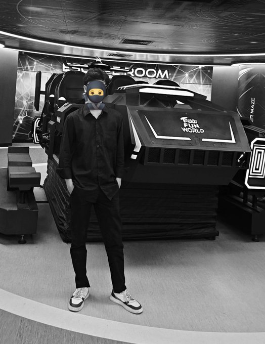 Black-and-white photo of a person in a dark shirt and pants standing in front of a 'Fun World' arcade racing game, wearing a cap with their face obscured by a monkey emoji.