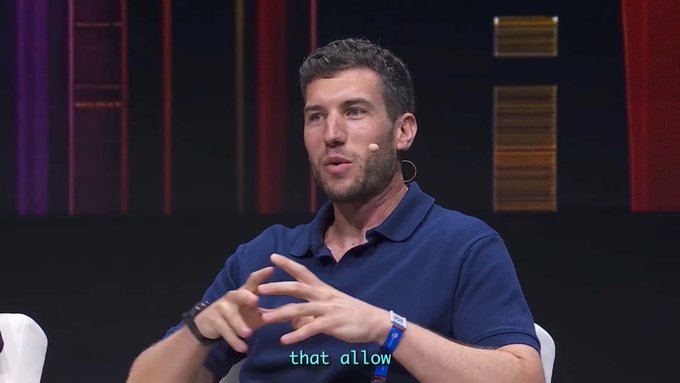 A man in a blue polo shirt with a wristband is gesturing while speaking at what appears to be a conference or panel discussion, with a small lapel microphone visible. The subtitle "that allow" appears at the bottom, relating to the post about protocols upgrading directly onto Sui and Walrus to bypass custom L1 overhead.