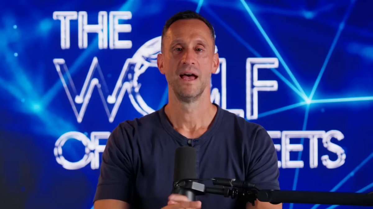 The image features a man wearing a blue shirt, sitting behind a microphone with his hands on the controls of a radio or podcast equipment. The background shows a large blue screen displaying the words "The Wolf of Bitcoin" along with some text about Bitcoin's value and market sentiment. This suggests that the man is likely discussing the current state of Bitcoin or its impact on the digital currency market, as indicated by the post.