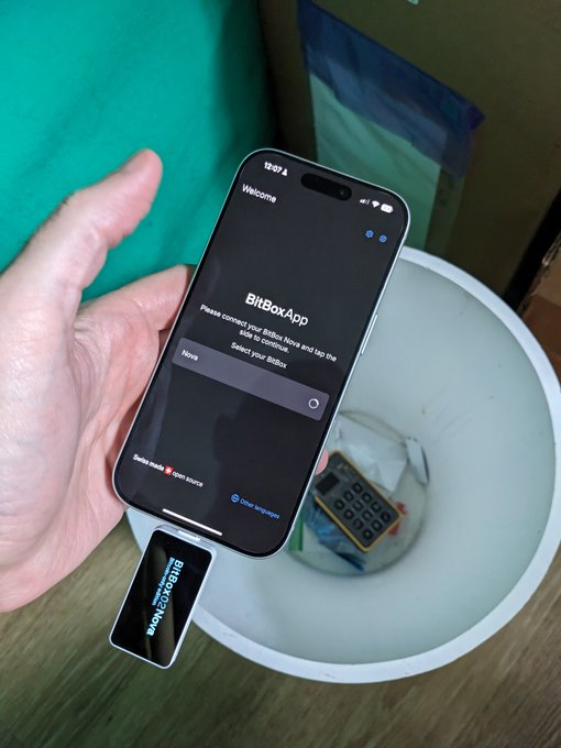 The image shows a hand holding an iPhone displaying the BitBoxApp setup screen above a white bucket containing a BitBox02 Nova hardware wallet, humorously framed as the user "throwing away" the other wallet's seed/backup to declare their loyalty to the BitBoxNova.