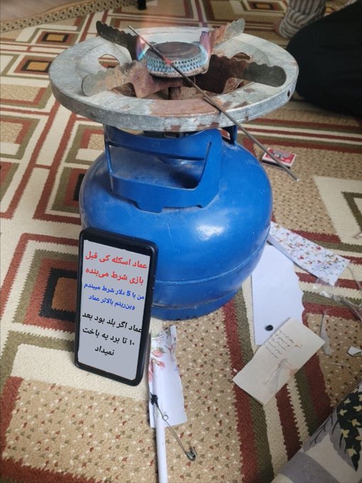 A blue gas cylinder sits on a patterned carpet with an empty wallet displayed on top alongside scattered price tags, while a phone shows Persian text from Emad Mousavi about his $25,000 loss on Polymarket betting markets, visually symbolizing his complete financial ruin from gambling.