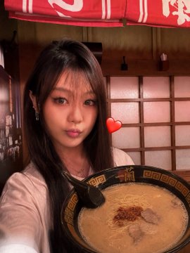 A selfie of a young woman with long dark hair taking a photo while holding a bowl of ramen (tonkotsu-style) with a black spoon, set in a restaurant interior with wooden sliding doors and a red noren curtain.