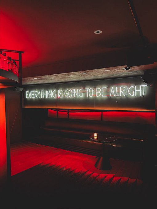 A dimly lit lounge area bathed in red light features a neon sign reading 'EVERYTHING IS GOING TO BE ALRIGHT' above a black leather booth and small table.