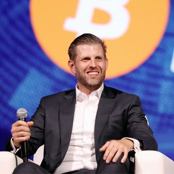 Eric Trump smiles while holding a microphone on stage, seemingly in connection with the news about American Bitcoin Corp's ($ABTC) increased Bitcoin holdings. The image likely depicts Eric Trump at an event related to the company or the cryptocurrency space.