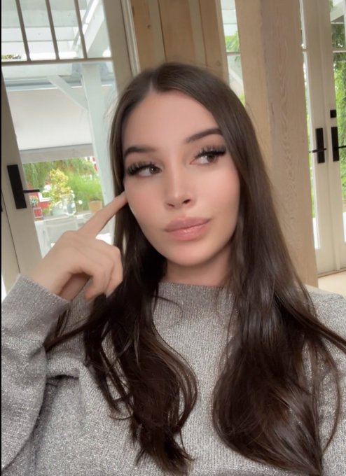 A young woman with long brown hair and makeup points to her temple in a selfie, wearing a gray sweater with a window and greenery visible behind her.