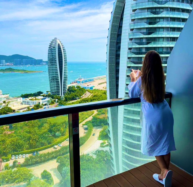 A woman in a white dress stands on a high-rise balcony, taking a photo of the scenic view that includes a distinctive curved building and the ocean.