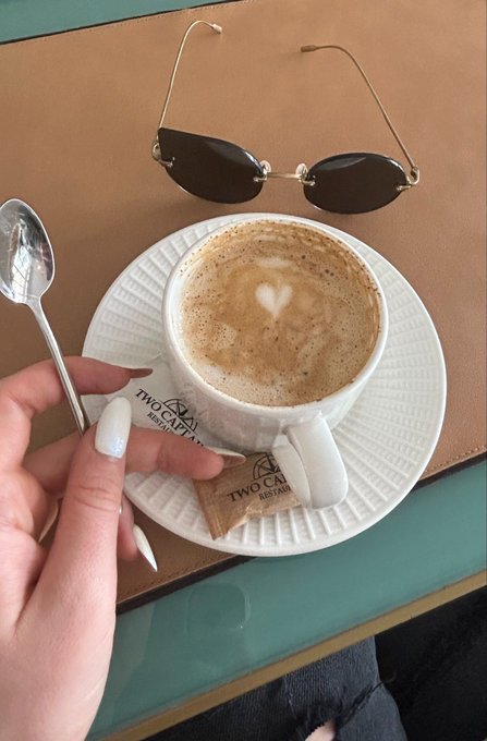 A hand holding a spoon beside a cup of coffee on a saucer from 'TWO CAPE RESTAURANT', with sunglasses resting on top of the saucer and the person sporting white French-tip nails.