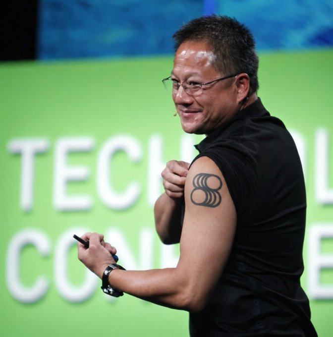 A smiling man wearing glasses and a black sleeveless shirt presents at a tech conference, showing a tattoo on his upper arm while holding a clicker.