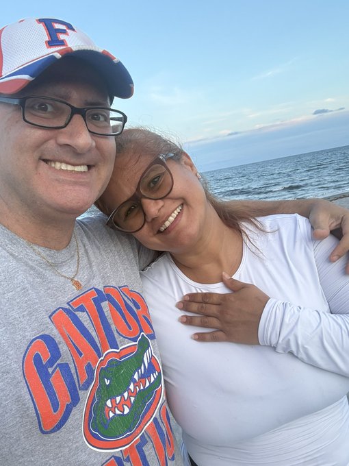 A selfie of a smiling couple at the beach; the man wears a Florida Gators shirt and a cap, while the woman in a white long-sleeve top rests her head on his shoulder.