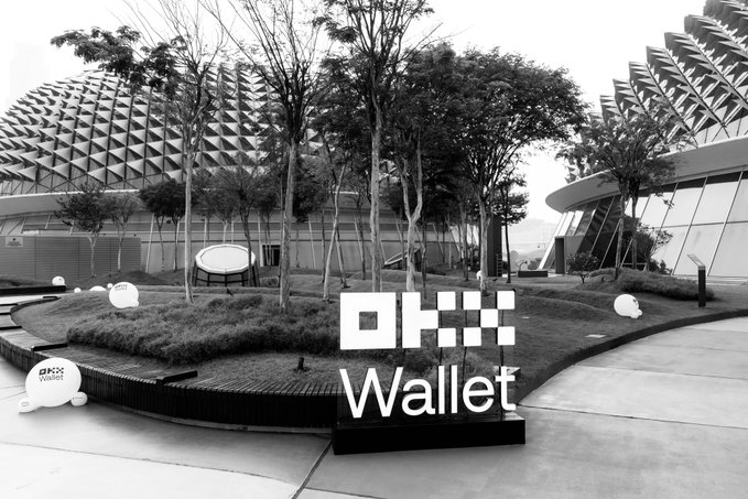 An outdoor event venue at night featuring the iconic spiky domes of the Esplanade Theatres in Singapore, with a large "OKX Wallet" sign prominently displayed in the foreground. The black-and-white scene, complete with decorative pig sculptures and lush landscaping, sets the stage for OKX's "Beyond" campaign event.