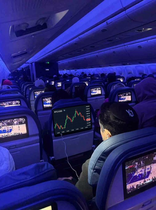 Inside a dimly lit airplane cabin with blue lighting, a passenger in a 'YO' cap watches a 'BTC/USD' candlestick chart on the seatback screen.