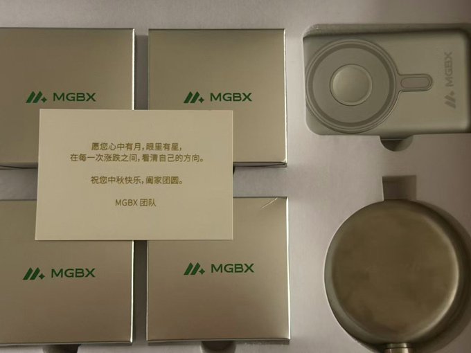 A Mid-Autumn Festival gift box arrangement from MGBX, featuring gold boxes branded with the MGBX logo, a handwritten card in Chinese wishing prosperity ('愿您心中有月，眼里有星'), and a metal water bottle and pot.