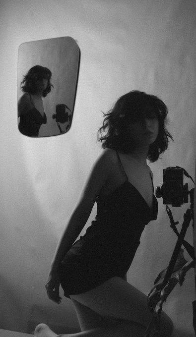 A black and white image features a woman in a black dress posing beside a vintage camera and a wall mirror, with her reflection visible; the accompanying text identifies the work as "Echoes of the self" 1/1 120 XTZ.